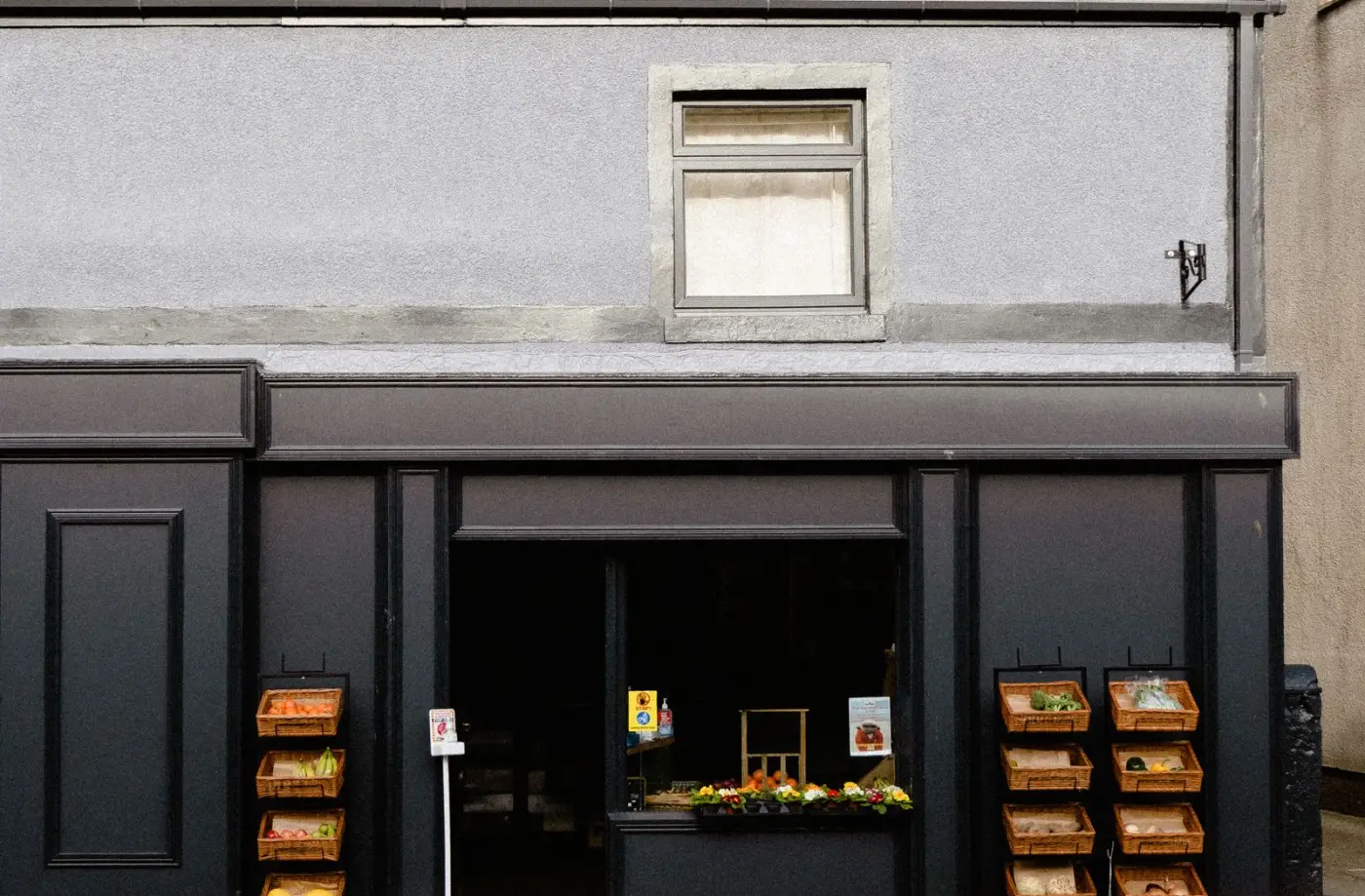A small greengrocer's shopfront seen straight-on from the street in daylight — a dark timber frontage with an open doorway and wicker baskets of fruit and vegetables to either side.
